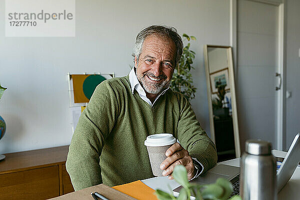 Lächelnder reifer Mann  der eine Einweg-Kaffeetasse hält und zu Hause sitzt