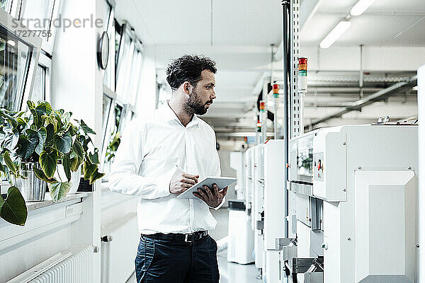 Geschäftsmann hält ein digitales Tablet in der Hand  während er sich Maschinen in der hellen Industrie ansieht