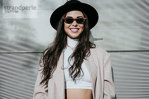 Lächelnde Frau mit Hut  die an einem sonnigen Tag an der Wand steht
