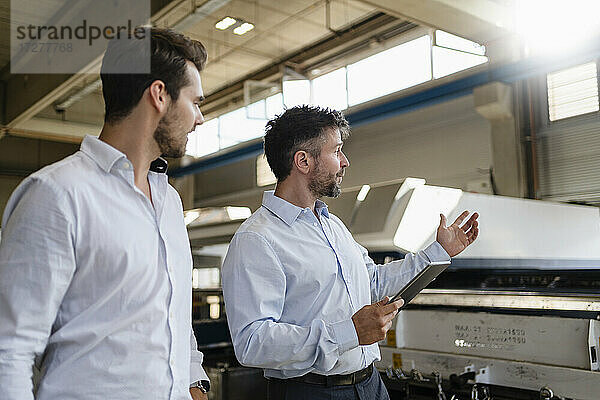 Reifer Geschäftsmann mit digitalem Tablet im Gespräch mit einem Kollegen  während er in einer Fabrik steht