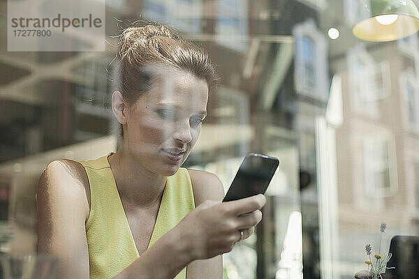 Lächelnde Geschäftsfrau  die ein Smartphone in einem Café benutzt  gesehen durch ein Glasfenster