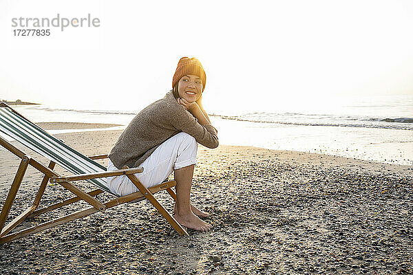 Lächelnde junge Frau  die auf einem Klappstuhl sitzt und bei Sonnenuntergang gegen den klaren Himmel auf den Strand blickt