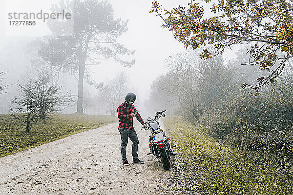 Mann benutzt sein Smartphone  während er auf einem unbefestigten Weg neben einem Motorrad steht
