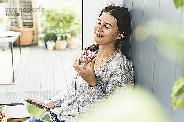 Frau mit geschlossenen Augen  die ein digitales Tablet und einen Donut hält  während sie zu Hause sitzt