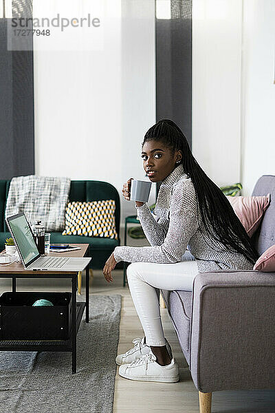 Unternehmerin trinkt Kaffee  während sie wegschaut  während sie zu Hause im Wohnzimmer sitzt