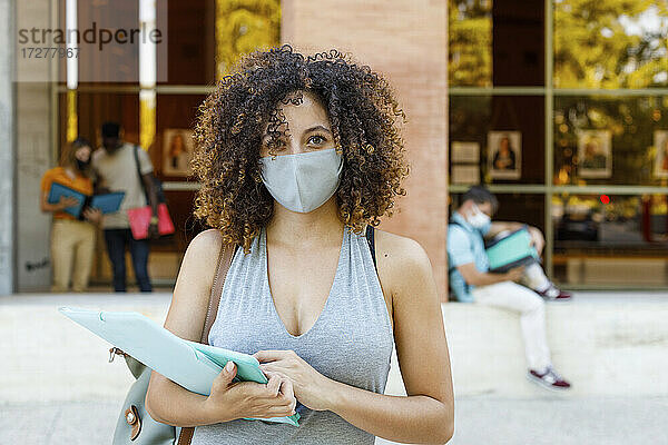 Junge Frau mit Dokumenten  die einen Gesichtsschutz trägt  während sie auf dem Universitätsgelände steht