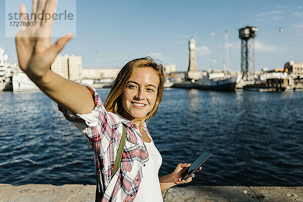 Frau  die am Meer steht und ihr Smartphone in der Hand hält  winkt an einem sonnigen Tag