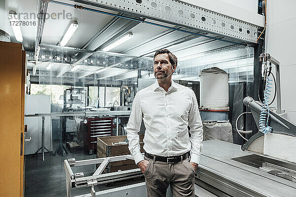 Nachdenklicher männlicher Ingenieur  der mit den Händen in den Taschen vor Maschinen in einer Fabrik steht