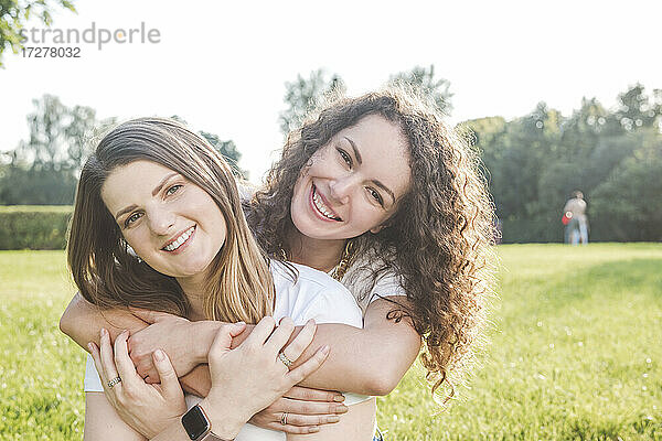 Lächelnde junge Frau umarmt ihre Freundin von hinten im Park an einem sonnigen Tag