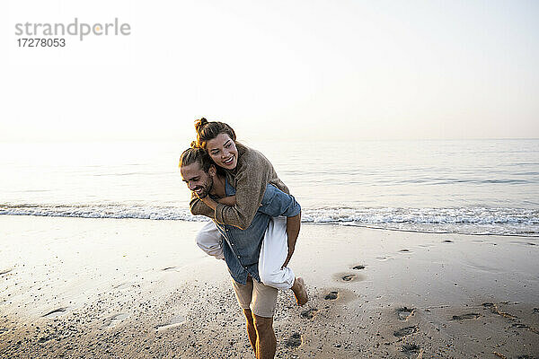 Glücklicher Mann  der seine Freundin huckepack nimmt  während er am Strand gegen den klaren Himmel bei Sonnenuntergang spazieren geht