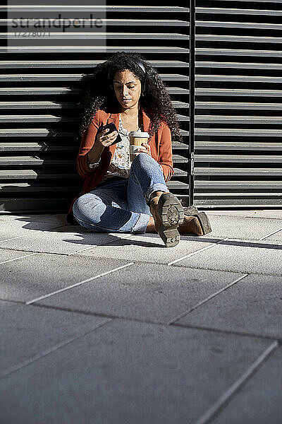 Frau  die ein Smartphone benutzt und eine Kaffeetasse hält  während sie gegen einen Fensterladen sitzt