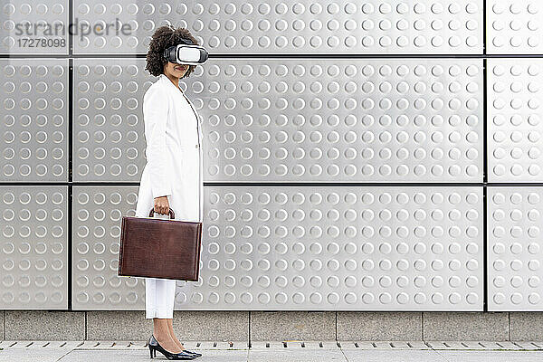 Geschäftsfrau mit Aktentasche  die eine Virtual-Reality-Brille benutzt  während sie vor einer silbernen Wand steht