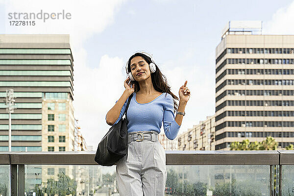 Junge Geschäftsfrau genießt Musik auf Kopfhörern  während sie sich auf einem Geländer gegen den Himmel lehnt