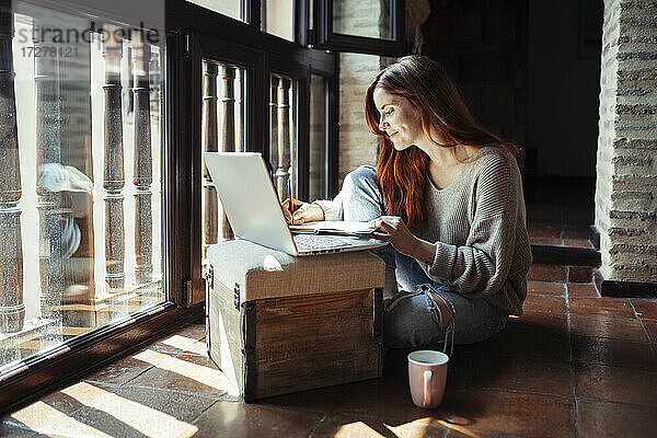 Lächelnde rothaarige Frau  die in ein Buch schreibt  während sie zu Hause einen Laptop benutzt