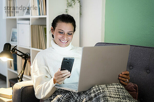 Lächelnde Frau benutzt Smartphone und Laptop  während sie zu Hause sitzt