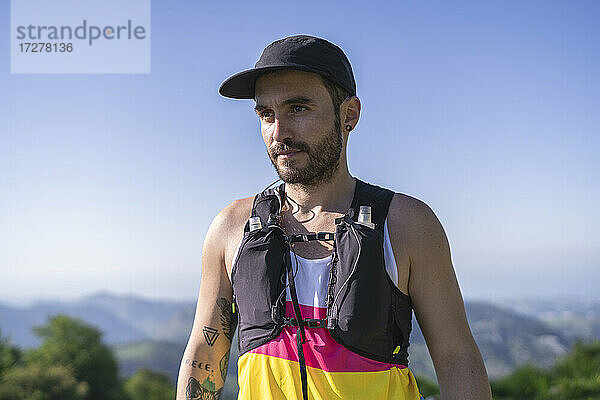 Selbstbewusster Sportler  der einen Flaschengurt trägt und die Aussicht bewundert  während er auf einem Berg gegen den klaren Himmel steht