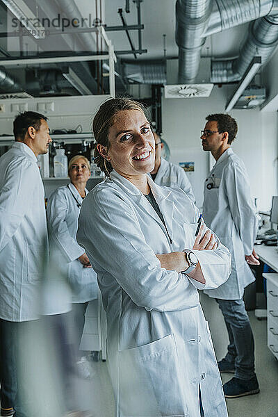 Junge Wissenschaftlerin lächelt  während sie mit verschränkten Armen und einem Mitarbeiter im Hintergrund im Labor steht
