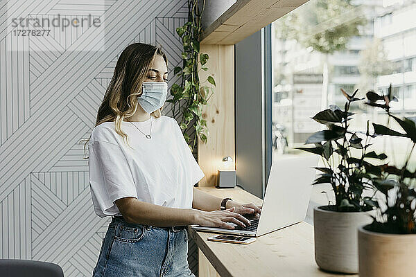 Frau mit Gesichtsschutzmaske  die einen Laptop benutzt  während sie in einem Café steht (Covid-19)