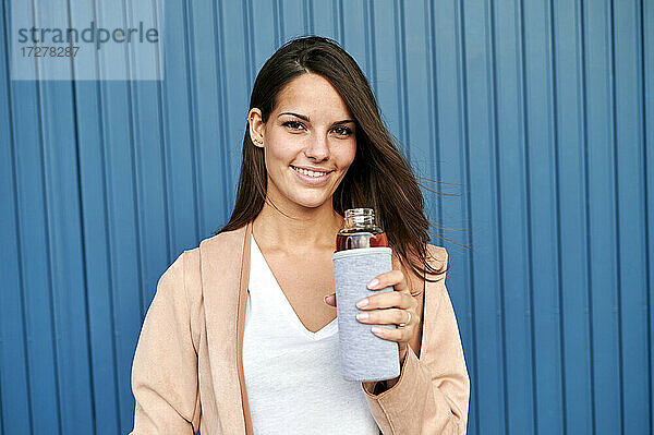 Junge Frau mit Mehrwegflasche  die an einer blauen Metallwand steht