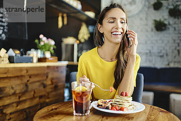 Lächelnde junge Frau  die eine Beere isst  während sie mit ihrem Smartphone am Tisch in einem Café spricht