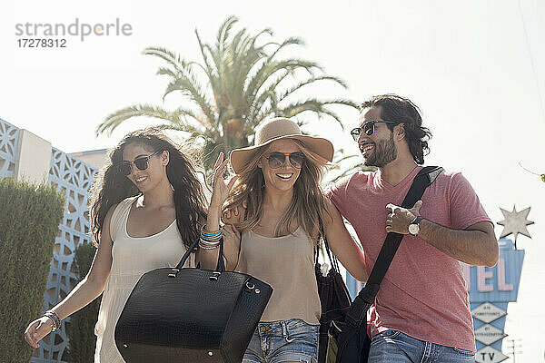 Lächelnde männliche und weibliche Freunde mit Sonnenbrillen gehen gegen den Himmel an einem sonnigen Tag