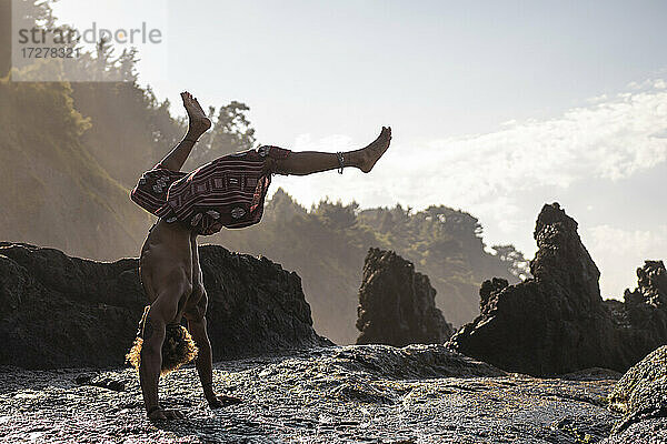 Junger Mann übt Handstand auf einer Felsformation am Strand gegen den Himmel
