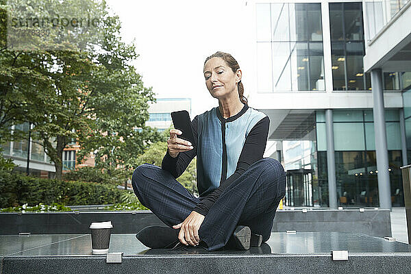 Geschäftsfrau  die ein Mobiltelefon benutzt  während sie auf einer Bank im Büropark sitzt