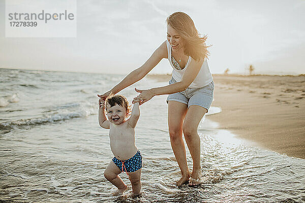 Fröhliche Mutter und Sohn genießen im Wasser am Strand an einem sonnigen Tag bei Sonnenuntergang