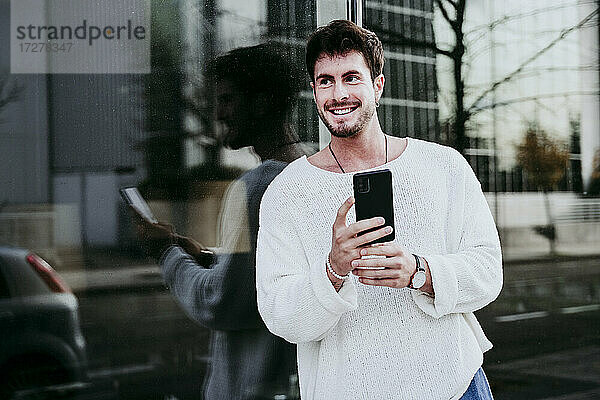 Lächelnder Mann mit Smartphone  der von einem Gebäude in der Stadt wegschaut
