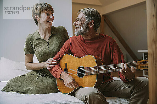 Reifer Mann spielt Gitarre  während die Frau ihn im Schlafzimmer auf dem Bett sitzend anschaut