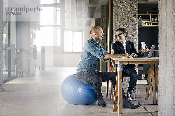 Geschäftsmann benutzt Laptop  während er neben einer Frau auf einem Fitnessball im Büro sitzt