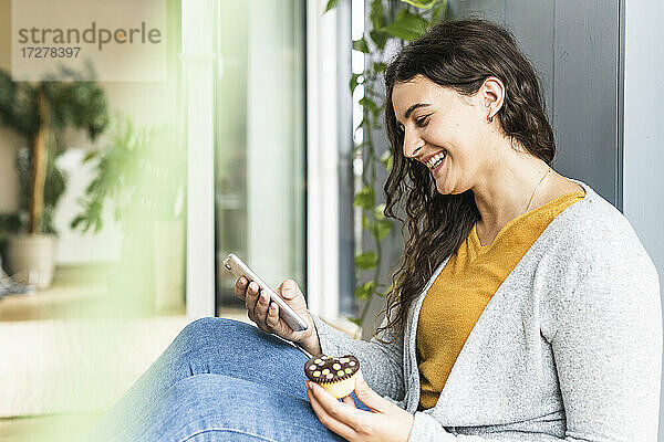 Lächelnde Frau mit Törtchen  die ihr Handy benutzt  während sie zu Hause sitzt