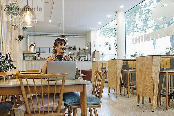 Lächelnde Unternehmerin  die eine Kaffeetasse hält und einen Laptop benutzt  während sie wegschaut und in einem Café sitzt