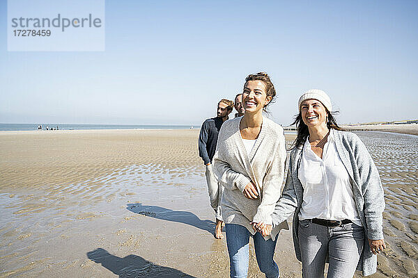 Lächelnde Frauen halten sich an den Händen  während sie mit Männern im Hintergrund am Strand spazieren gehen