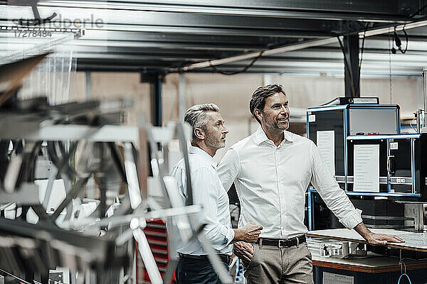 Ein reifer Kollege diskutiert mit einem lächelnden Manager  während er an einer Fertigungsmaschine in einer Fabrik steht