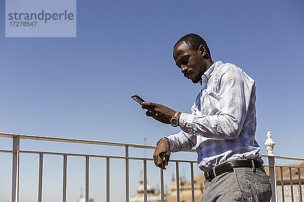 Männlicher Unternehmer  der sein Smartphone benutzt  während er sich gegen den klaren blauen Himmel auf ein Geländer stützt