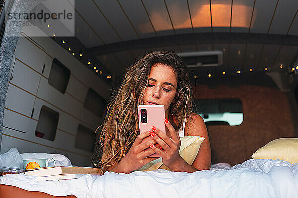 Schöne Frau  die ein Mobiltelefon benutzt  während sie bei Sonnenuntergang am Strand im Wohnmobil liegt