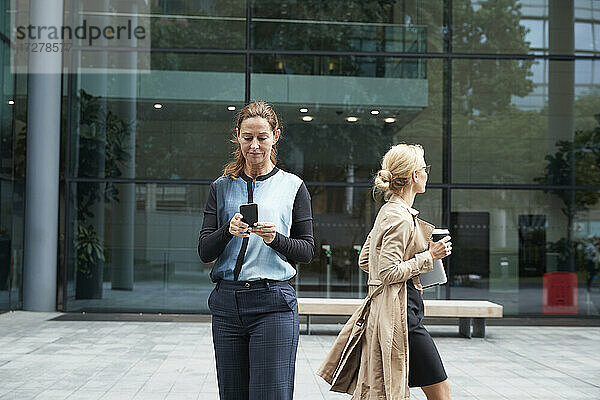 Geschäftsfrau  die ein Mobiltelefon benutzt  mit einem Kollegen  der nach der Arbeit geht  im Hintergrund