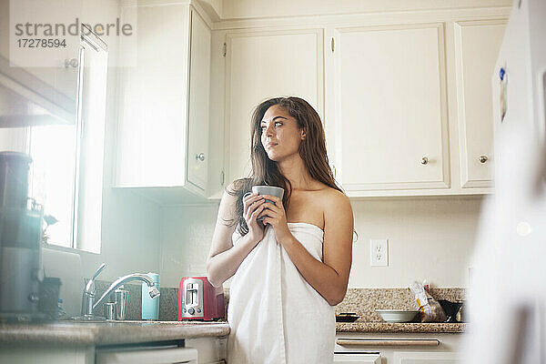 Nachdenkliche Frau  die ein Handtuch trägt und eine Kaffeetasse hält  während sie in der Küche zu Hause steht