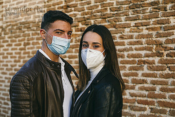 Junges Paar mit Gesichtsmaske  das zusammen an einer Backsteinmauer steht