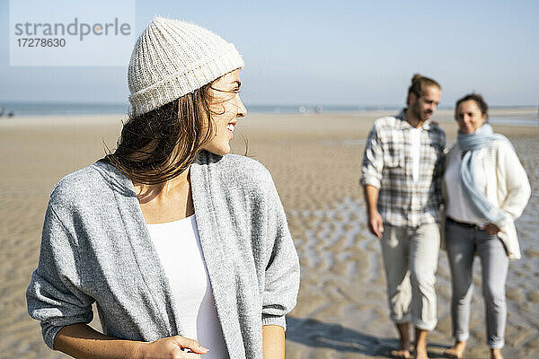 Junge Frau schaut über die Schulter zu einem Mann und einer Mutter  die im Hintergrund am Strand spazieren gehen