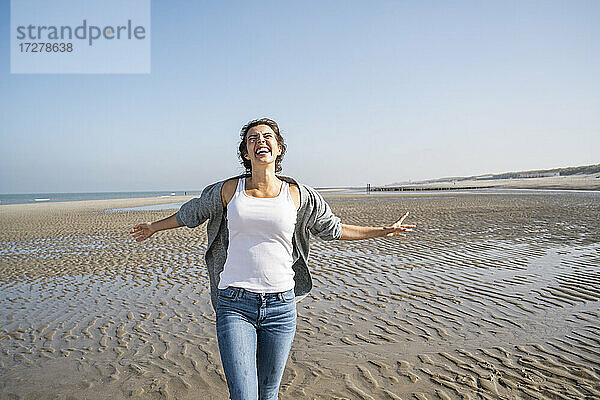 Fröhliche junge Frau mit ausgestreckten Armen  die am Strand gegen den klaren Himmel spazieren geht