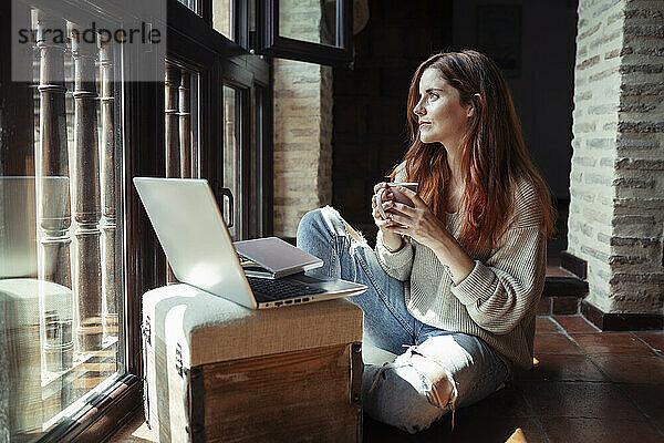 Junge Frau trinkt Kaffee  während sie zu Hause am Laptop arbeitet