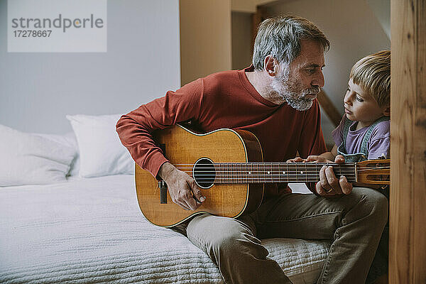 Vater spielt Gitarre vor seinem Sohn  während er zu Hause auf dem Bett sitzt