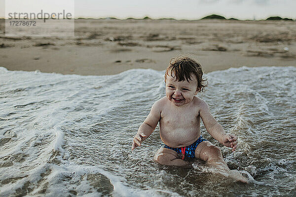 Fröhlicher kleiner Junge spielt im Wasser am Strand bei Sonnenuntergang
