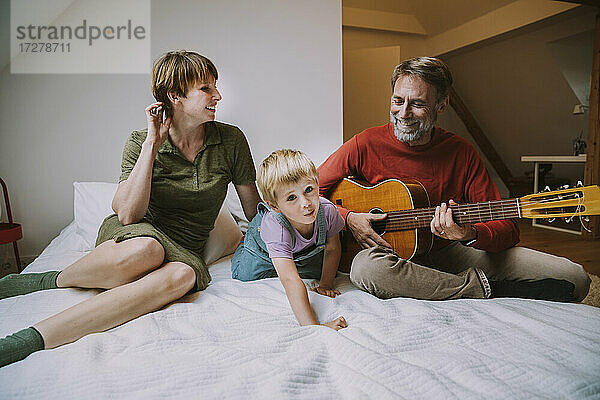 Glücklicher Vater spielt Gitarre  während Mutter und Sohn zu Hause auf dem Bett sitzen