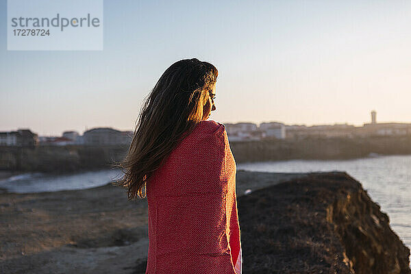 Junge Frau mit bedecktem Schal  die am Strand stehend die Aussicht betrachtet