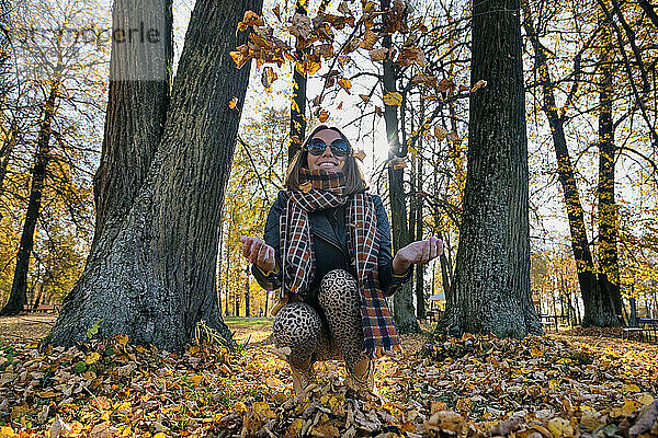 Fröhliche Frau  die trockene Herbstblätter wirft  während sie sich im Park an Bäume kauert