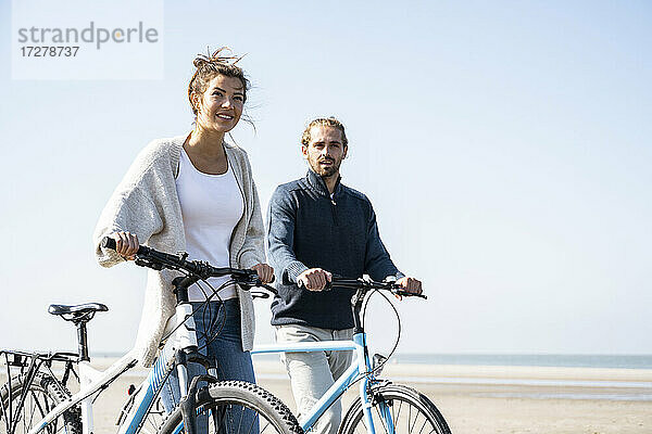 Junges Paar zu Fuß mit Fahrrädern am Strand gegen klaren Himmel an einem sonnigen Tag