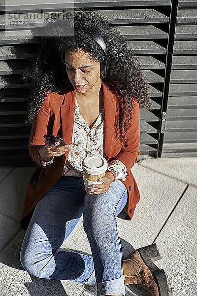 Junge Frau  die ein Smartphone benutzt und eine Kaffeetasse hält  während sie auf einem Fußweg sitzt